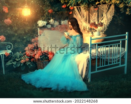 Artwork Fairy tale princess sits on vintage bed. Fantasy woman holding cup in hands drinking tea. Long wave hair. Blue vintage luxury dress. Backdrop blooming flowers, old lamp. Fantasy woman queen