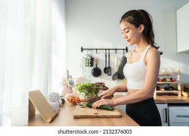 Asaian attractive woman wear sport clothes cook green salad in kitchen. Young beautiful girl feeling happy and enjoy eating vegetables healthy foods to diet and lose weight for health care in house. - Shutterstock ID 2132376003