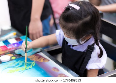 Asain Little Child Girl Wearing A White Cloth Mask Is Learning To Draw Art With Watercolor And Brush. Adorable Kid Wore A Black Apron Sitting To Draw Pictures. Child 3 Years Old.