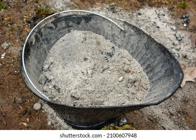 A ash bucket full of wood ashes sitting outside on the ground- Garden fertilizer- the remains of burnt firewood- A black ash bucket full of wood ash