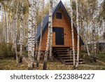 A-shaped frame house in Scandinavian style. The landscape. Quiet rest. The concept of glamping and chalet rentals. A wooden house with a veranda in nature around an autumn birch forest.