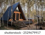 A-shaped frame house in Scandinavian style. The landscape. Quiet rest. The concept of glamping and chalet rentals. A wooden house with a veranda in nature around an autumn birch forest.