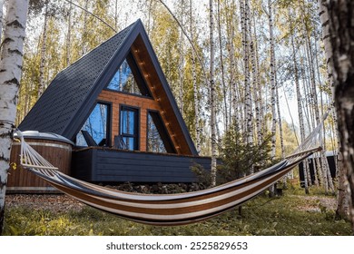 A-shaped Scandinavian style frame house and hammock. The landscape. Quiet rest. The concept of glamping and chalet rentals. A wooden house with a veranda in an autumn birch forest. - Powered by Shutterstock