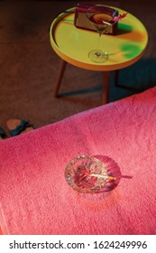 Ashtray With Burning Cigarette On Sunbed With Vintage 1950s Ladies Shoes And Yellow Table With Cocktail And Radio Next To It.