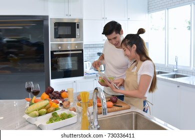 Asia Couple Is Looking At Each Other And Smiling While Cooking In Kitchen At Home. Man Is Mixing Salad.