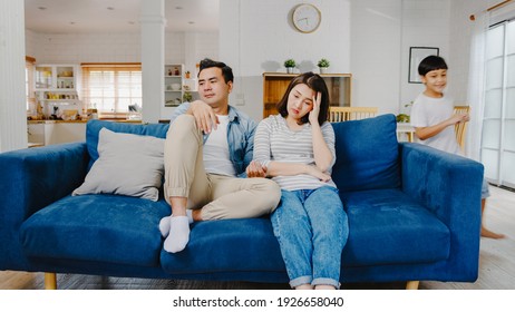 Asia Family Dad And Mom Sit On Couch Feels Annoyed Exhausted While Daughter And Son Have Fun Shouting Run Around Sofa In Living Room At Home. Social Distancing, Quarantine For Corona Virus Prevention.