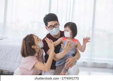 Asia Family In Medical Masks On The Face Looks At The Camera While Standing In The Living Room At Home To Prevent PM2.5 Dust, Smog, Air Pollution And COVID-19. Healthcare Concept.