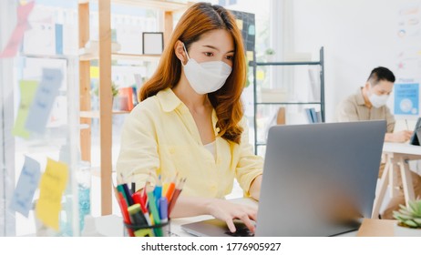 Asia Female Wear Face Mask For Social Distancing In New Normal Situation For Virus Prevention While Using Laptop And Separated By Acrylic Partition Stand In Office. Life And Work After Corona Virus.