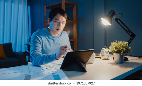 Asia freelance businessman focus work pen write on tablet computer busy with full of graph paperwork on desk in living room at home overtime at night, Work from home during COVID-19 pandemic concept. - Shutterstock ID 1971857927