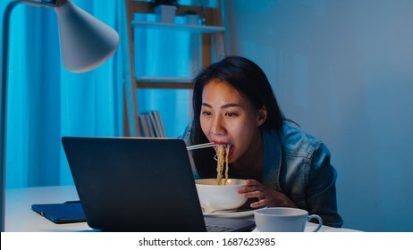 Asia Freelance Smart Business Women Eating Instant Noodles While Working On Laptop In Living Room At Home At Night. Happy Young Asian Girl Sitting On Desk Work Overtime, Enjoy Relax Time.