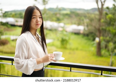An Asia girl in white shirt is standing on balcony and holding a white cup with blur background and copy space from Thailand.