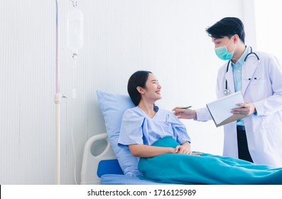 Asia Man Doctor Wearing Gown Coat And Stethoscope Surgical Mask Holding Noteplate.Physician Standing Adivises Asia Smiley Woman Patients Lying On Bed In Hospital Clinic Copy Space