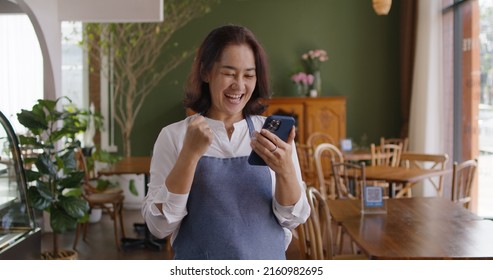 Asia mature adult woman people wear apron small cafe receive e-mail text good news SME lending service solution win seller store in social media app. Shock smile laugh face in sale order reopen shop.