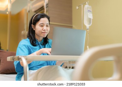 Asia Patient Woman Sitting On The Bed With Saline Solution In Hospital Ward Using Laptop, Computer And Check Email. Working In Clinic. Busy Life Of Employee During Sick. Healthcare And Medical Concept