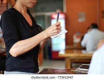 The Asia waitress writing the order from customer. / selective focus on hand and blue pen of waitress / background with blur customer in the restaurant . / Taking a note to the paper.