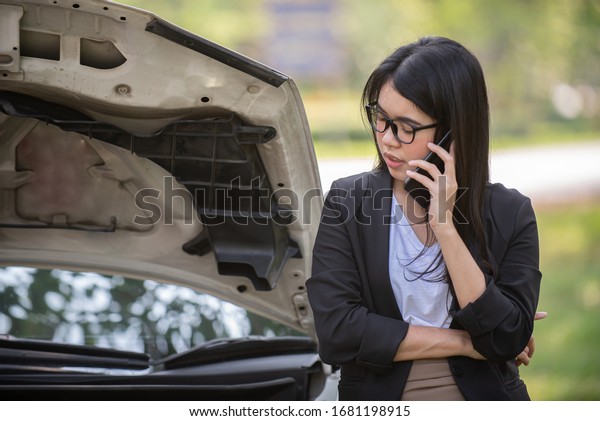 Asia young woman is standing near her\
broken car. She needs for help. The girl is talking on the mobile\
phone and smiling. The bonnet of her car is\
open
