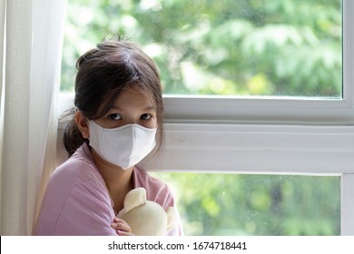 Asian 6 Or 7 Years Old Kid Wearing Medical Mask.Little Girl Standing By The Window And Looking Outside.She Look Sad,bored.She May Sick  Or Quarantine From The Coronavirus Disease 19 (COVID-19). 