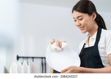 Asian active cleaning service woman worker cleaning in kitchen at home. Beautiful young girl housekeeper cleaner feel happy and wipes dishes plates after washing for housekeeping housework or chores.