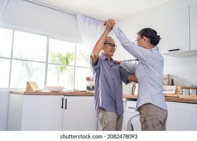 Asian Active Senior Couple Enjoy Dancing Together In Kitchen At Home. Elderly Older Grandfather And Grandmother Smiling Feeling In Love And Enjoy Relationship And Activity In House After Retirement.