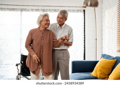 Asian active senior man support elderly woman from wheelchair to sofa. Happy family, Beautiful mature older couple doing physical therapy after retirement for health together in living room at home. - Powered by Shutterstock