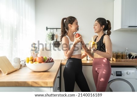 Asian active two women sibling in sportswear eat an apple in kitchen. Young beautiful girl sister feeling happy and enjoy eating fruits healthy foods to diet and lose weight for health care in house.