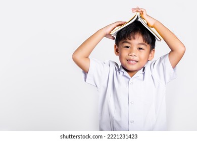 Asian Adorable Toddler Smiling Happy Wearing Student Thai Uniform Red Pants Stand Holding Book Over Head Like Roof In Studio Shot Isolated On White Background, Portrait Little Children Boy Preschool