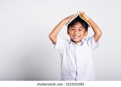 Asian Adorable Toddler Smiling Happy Wearing Student Thai Uniform Red Pants Stand Holding Book Over Head Like Roof In Studio Shot Isolated On White Background, Portrait Little Children Boy Preschool