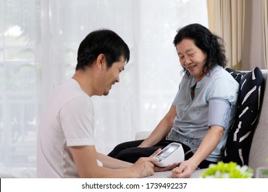 Asian Adult Son Helping His Elderly Mother To Check Blood Pressure Monitor For The Hypertension Disease Care At Home, Concept Of Healthcare And Lifestyle Of Elderly People In The Family.