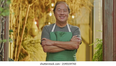 An Asian American Business Owner Wearing Apron And Crossing Arms Smiling. Portrait Of An Asian Middle Aged Businessman Owner Of Small Local Store With Arms Crossed Looking At Camera