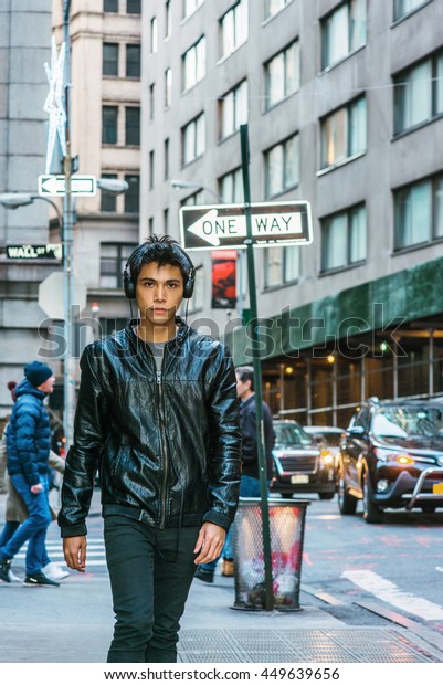 Asian American college student traveling in New
York. Wearing black leather jacket, headphone on head, young guy
walking on narrow street with high buildings. Cars, people on
background. City Life.
