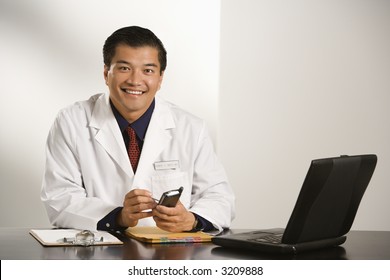 Asian American Male Doctor Sitting At Desk With Charts And Laptop Computer Using Pda.