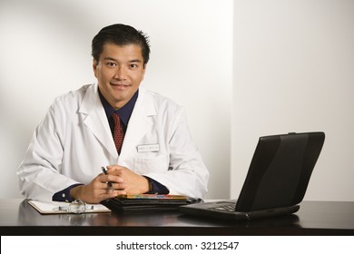 Asian American Male Doctor Sitting At Desk With Charts And Laptop Computer Looking At Viewer.