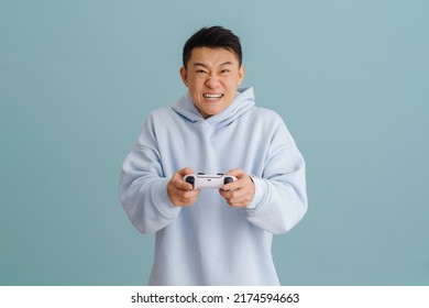 Asian angry man grimacing while playing video game with joystick isolated over blue background - Shutterstock ID 2174594663