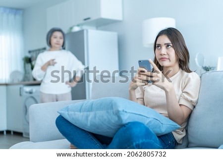 Asian angry senior mature woman shouting at stubborn fussy daughter. Young female sit on sofa use mobile phone chat, disobedient rebellious and ignore annoyed mother. Family problem in house concept.