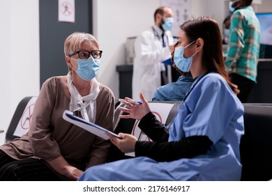 Asian Assistant Consulting Old Woman In Waiting Room, Talking About Virus Prevention And Healthcare At Hospital Reception. Elderly Patient And Medical Assistant Chatting During Covid 19 Pandemic.