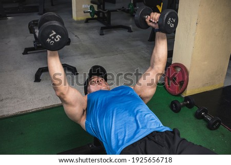 An asian athlete does a set of dumbbell flys on a flat bench, lowering the arms until inline with chest. Training pecs and upper body at the gym.