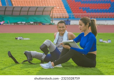 Asian athlete with prosthetics and his female trainer sit on a lawn, planning the next day's speed running practice at the stadium - Powered by Shutterstock