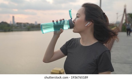 Asian Athlete Woman In Green Nature Tree Park And Lake In Evening Sunset Feeling Thirsty Drink Water From Bottle. Energy Drinks Refreshment Hydration From Tired And Exhausted. Workout And Drink Water.