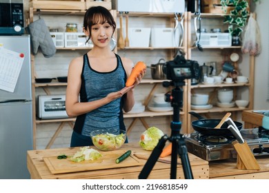 Asian Athletic Woman Holding A Carrot And Talking To Digital Camera On Tripod Is Showing The Ingredient While Vlogging About Making Salad In The Kitchen At Home