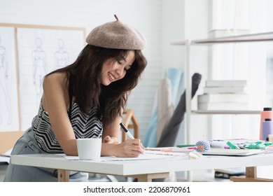 Asian Attractive 30s Woman Fashion Designer Or Dressmaker Drawing Sketch Of New Collection Clothes On Paper And Smiling While Working With Tablet And Tailoring Equipments On Desk At Workplace Studio