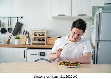 Asian Attractive Handsome Male Eating Green Salad In Kitchen At Home. Active Young Man Sit On Table, Feel Happy And Enjoy Vegetables Healthy Foods And Clean Water For Breakfast To Diet For Health Care