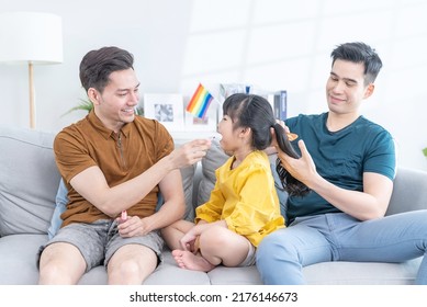 Asian Attractive LGBTQ Gay Family Spending Time With Girl Kid Daughter. Handsome Male Couple Sit On Sofa In Living Room, Take Care And Comb Little Adorable Child Hair Enjoy Parenting Activity At Home.