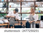 Asian attractive male barista or waiter working in coffeehouse shop. Young handsome entrepreneur man in apron teaching waitress grinding roasted beans making coffee on counter at urban restaurant cafe