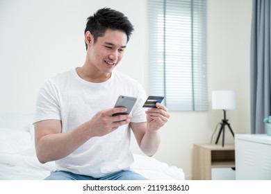 Asian Attractive Male Enjoy Shopping Online On Bed At Home In Morning. Handsome Man Sitting In Bedroom, Holding Credit Card To Make E Bank Online Payment After Use Phone Purchase In Web Store In House