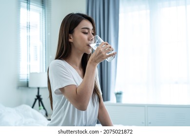 Asian Attractive Woman Sit On Bed, Drink A Glass Of Water In Bedroom. Young Thirsty Beautiful Girl Holding Clean Mineral Natural In Cup After Wake Up And Sipping In Morning For Health Care In House.