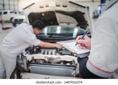 Asian Auto Mechanic Is Checking The Engine And Analyzing The Car Engine Problem And Writing To Clipboard. Checklist For Machine Repair, Car Service And Maintenance.