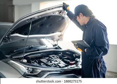 Asian auto mechanic holding digital tablet checking car engine under bonnet hood in auto service garage. Mechanical maintenance engineer working in automotive industry. Automobile servicing and repair