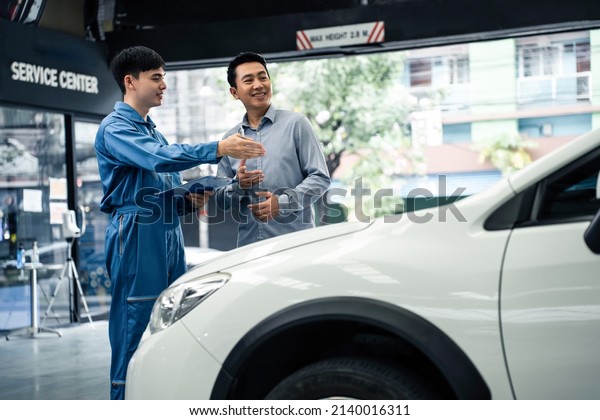 Asian automotive mechanic explain car condition to\
client in garage. Vehicle service manager male work in mechanics\
workshop point at vehicle part to customer man for maintenance and\
repair car engine
