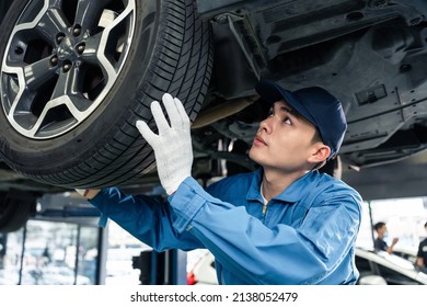 Asian automotive mechanic repairman look under car condition in garage. Vehicle service guy worker doing check or mend car wheel and work in mechanics workshop with confidence to repair car engine car