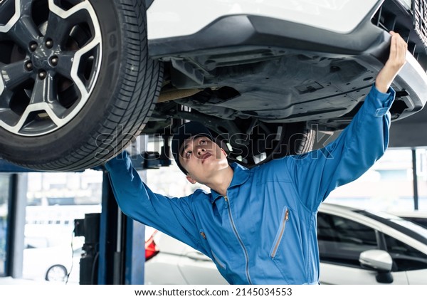 Asian automotive mechanic repairman look under car\
condition in garage. Vehicle service guy worker doing check or mend\
car wheel and work in mechanics workshop with confidence to repair\
car engine car